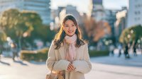 Young Asian businesswoman in beige coat standing confidently in modern urban plaza during golden hour with city skyline background for corporate lifestyle.