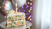 Colorful funfetti birthday cake slice with vanilla frosting and lit candle on glass plate against festive bokeh lights background for celebration.