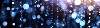 Glowing blue bokeh lights and sparkling particles on dark background create magical atmosphere for festive designs and holiday decorations.