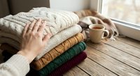 Woman's hands touching stack of cozy knitted sweaters in warm autumn colors on wooden table near window with coffee cup creating hygge atmosphere.
