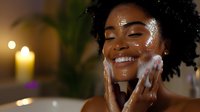 Young African American woman with glowing skin applying facial cleanser, smiling with eyes closed in warm candlelit spa setting, showcasing skincare and wellness routine.