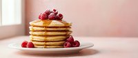Stack of golden pancakes topped with fresh raspberries and maple syrup on white plate in bright kitchen setting for breakfast and brunch concepts.