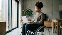 Woman with dark curly hair, medium skin tone, using laptop while sitting in wheelchair in modern home office with natural light and plants.