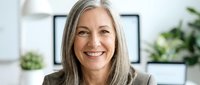 Mature woman with long gray hair and fair complexion smiling at camera in bright office environment with green plants and modern decor, representing confidence and professionalism.