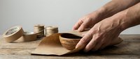 Male hands crafting with natural brown paper tape rolls on wooden surface for packaging and shipping supplies in workshop setting.
