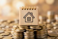 Small notepad with house icon sits on stacks of golden coins representing real estate investment, mortgage planning, property savings and home financing.