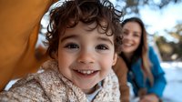 Young curly-haired child smiling at camera with genuine joy while parent watches from background in winter setting, capturing authentic family moment and emotional connection.