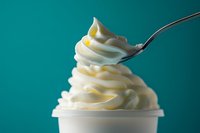 Creamy soft serve vanilla ice cream swirled high in white paper cup with metal spoon against turquoise background, macro food photography highlighting smooth texture.