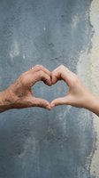 Two hands forming heart shape showing intergenerational love and connection between young and elderly person against textured concrete background.
