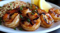 Grilled jumbo shrimp with caramelized glaze served on white plate alongside brown rice pilaf and lemon wedge garnish, topped with fresh herbs.