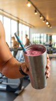 Person holding stainless steel tumbler with pink smoothie and blue straw in modern cafe or gym setting with blurred background and natural light.