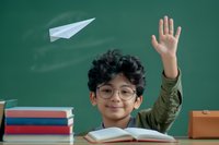 Asian elementary school student raising hand in classroom with paper airplane flying near chalkboard, books on desk, wearing glasses and green jacket.
