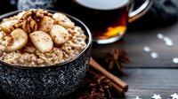 Creamy oatmeal topped with sliced bananas, walnuts and cinnamon in black ceramic bowl. Hot tea, star anise and cinnamon sticks on dark wooden background.