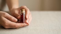 Hands holding small glass bottle with red liquid and cork stopper on neutral surface, suitable for wellness, aromatherapy, and product photography.