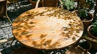 Wooden round table with dappled sunlight shadows on outdoor patio, surrounded by wicker chairs and potted plants, perfect for garden dining and exterior furniture styling.