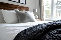 Cozy modern bedroom with white bedding, gray textured pillows and chunky knit black throw blanket against rustic wooden headboard, bright window view of pine trees.