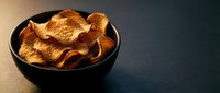 Crispy golden potato chips in a dark ceramic bowl on a moody black surface, copy space for food and snack advertising.