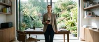 Middle-aged woman with light skin tone, wearing a beige blazer and black pants, standing in a modern home office with wooden desk, shelving, and large windows overlooking green garden landscape.