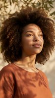 Young African American woman with natural curly afro hair in warm sunlight wearing orange top with tropical palm shadows creating artistic lighting patterns.