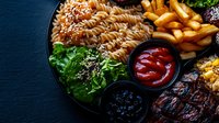Grilled steak dinner plate with fusilli pasta, french fries, fresh green salad, and assorted dipping sauces on dark background, close up view of restaurant meal.