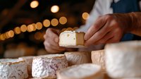 Artisanal cheese selection on rustic wooden board with hands serving fresh soft cheese wheel, atmospheric bokeh lights create cozy restaurant ambiance.