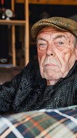 Senior Caucasian man with weathered facial features and blue eyes wearing a tan flat cap and dark herringbone jacket, seated indoors with plaid fabric visible, suitable for aging, lifestyle, and portrait concepts.