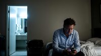 Depressed middle-aged Caucasian businessman sitting on bed edge using smartphone in dark hotel room with city view through bathroom door.