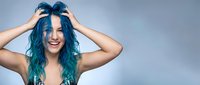 Young woman with vibrant blue-green hair laughing joyfully while running hands through colorful locks against soft gradient background for beauty campaigns.