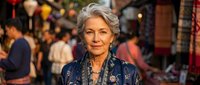 Elegant senior woman with silver hair in traditional blue clothing at vibrant marketplace. Mature female shopper exploring colorful bazaar stalls with warm lighting.