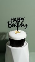 Elegant birthday cupcake with white fondant, gold leaf accent, and black Happy Birthday topper on sage green background for celebration.