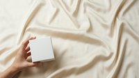 Hand holding blank white box on beige fabric background with soft shadows, suitable for product packaging mockups, branding presentations, and retail advertising campaigns.