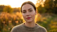 Young Caucasian woman in beige sweater smiling warmly at camera during golden hour sunset with autumn foliage background for lifestyle content.