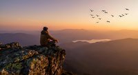 Hiker sitting on mountain cliff edge watching sunset over layered peaks with birds flying across golden sky for outdoor adventure and travel concepts.
