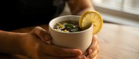 Hands holding warm bowl of herbal tea with lemon slice and fresh herbs, comfort beverage for wellness and relaxation.