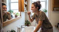 Young woman brushing teeth in modern bathroom mirror reflection showing daily morning hygiene routine self-care wellness lifestyle habits.