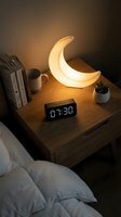 Cozy bedroom nightstand with glowing crescent moon lamp, digital clock, books and coffee mug creating warm ambient lighting for relaxation and sleep.