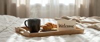 Wooden breakfast tray on bed with steaming black coffee cup, fresh croissant, and welcome card in cozy sunlit bedroom interior.