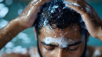 Young man washing hair with shampoo foam in shower, water drops on face and wet hair, eyes closed, self care and hygiene concept against turquoise background.