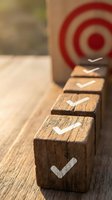 Wooden blocks with white checkmarks aligned in row on desk with red target bullseye in background, representing goal achievement, success milestones, and business progress.