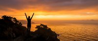 Person celebrating success with raised arms on cliff overlooking ocean at golden sunset, freedom and achievement concept for motivational content.