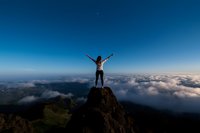 Young woman with raised arms standing on mountain peak above clouds at sunrise, expressing freedom and achievement against dramatic sky and landscape.