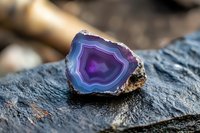 Purple and blue agate crystal slice with concentric layers and raw edges displayed on dark stone surface in natural lighting, showcasing mineral's natural banding pattern.