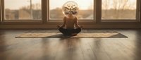 Young woman practicing meditation in lotus position on yoga mat in sunlit room with large windows during golden hour for mindfulness and wellness routine.