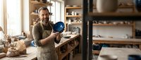 Middle-aged male artisan with beard, wearing apron, holding blue ceramic bowl in pottery studio workshop with wooden shelves and clay materials.