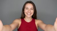 Young Caucasian woman in red tank top taking selfie with outstretched arms, smiling at camera against gray background for social media content.
