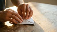 Close-up of hands folding white paper on wooden surface in natural sunlight, suitable for origami, crafts, DIY projects and creative activities.