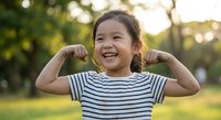 Happy Asian girl child flexing muscles outdoors showing strength and confidence in striped shirt during golden hour sunset for health and fitness concepts.