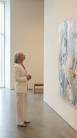 Mature Caucasian woman in white suit admiring abstract blue artwork in modern gallery space with natural light and minimalist design.