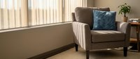 Cozy beige armchair with blue cushion in sunlit living room corner near window with natural light streaming through blinds creating warm atmosphere.