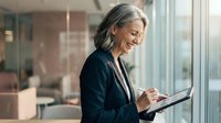Mature businesswoman with gray hair using digital tablet in modern office, smiling while working on technology device near window with natural light.